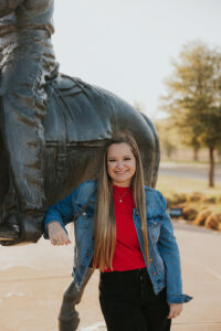 Marketing manager Scout in front of a horse statue wearing a red shirt, black pants, and jean jacket. She has her long brown hair down in front of her shoulders.