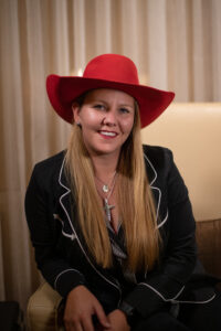 Headshot image of Natalie McFarland wearing a black suit, red hat, and long blonde hair in front.